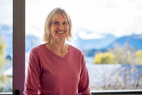 Catherine Syme, public speaking coach, smiling indoors with a view of Wanaka’s mountains through the window.